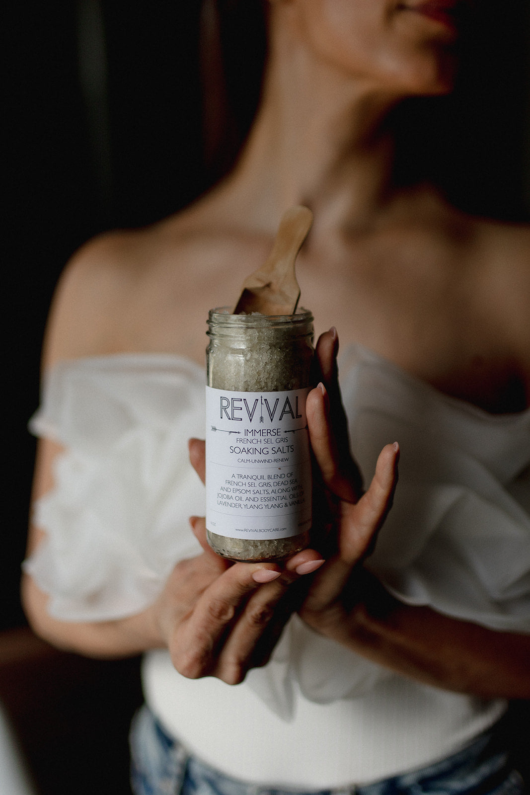 Person holding a jar of 'Revival' shower salts with a wooden scoop.