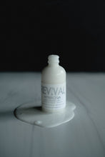 A white bottle with a label reading 'REVIVAL', Ambrosia, placed on a surface with a small amount of liquid around it, against a dark background.
