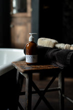 A bottle of Reviva botanical body lotion on a rustic wooden stool next to a towel and a dry brush, with a blurred background.