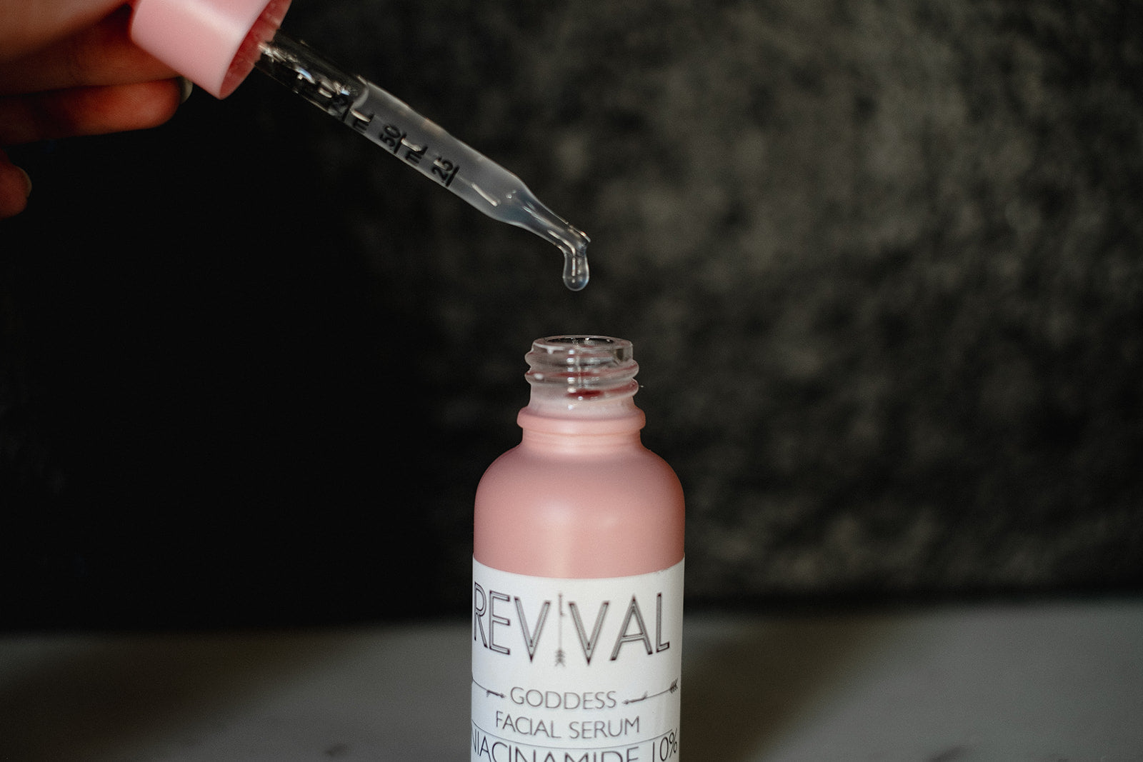 Pink facial serum bottle with dropper being used against a dark background
