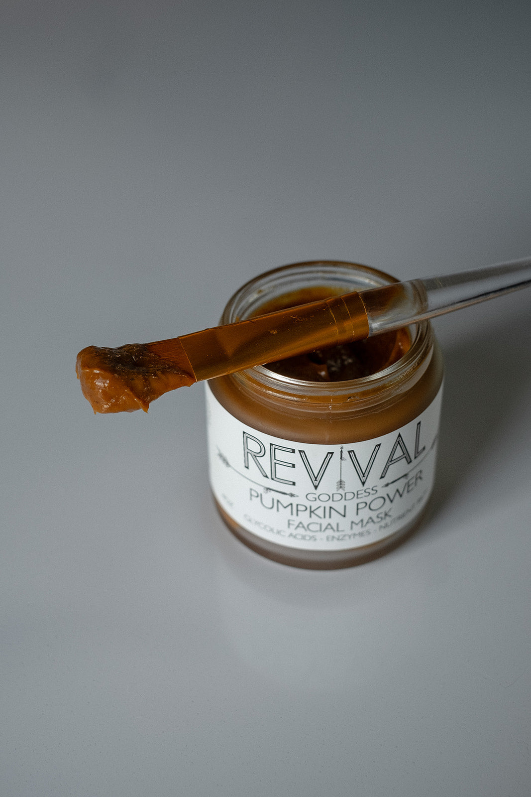 Jar of 'Revival' pumpkin power face mask with a applicator on a gray background