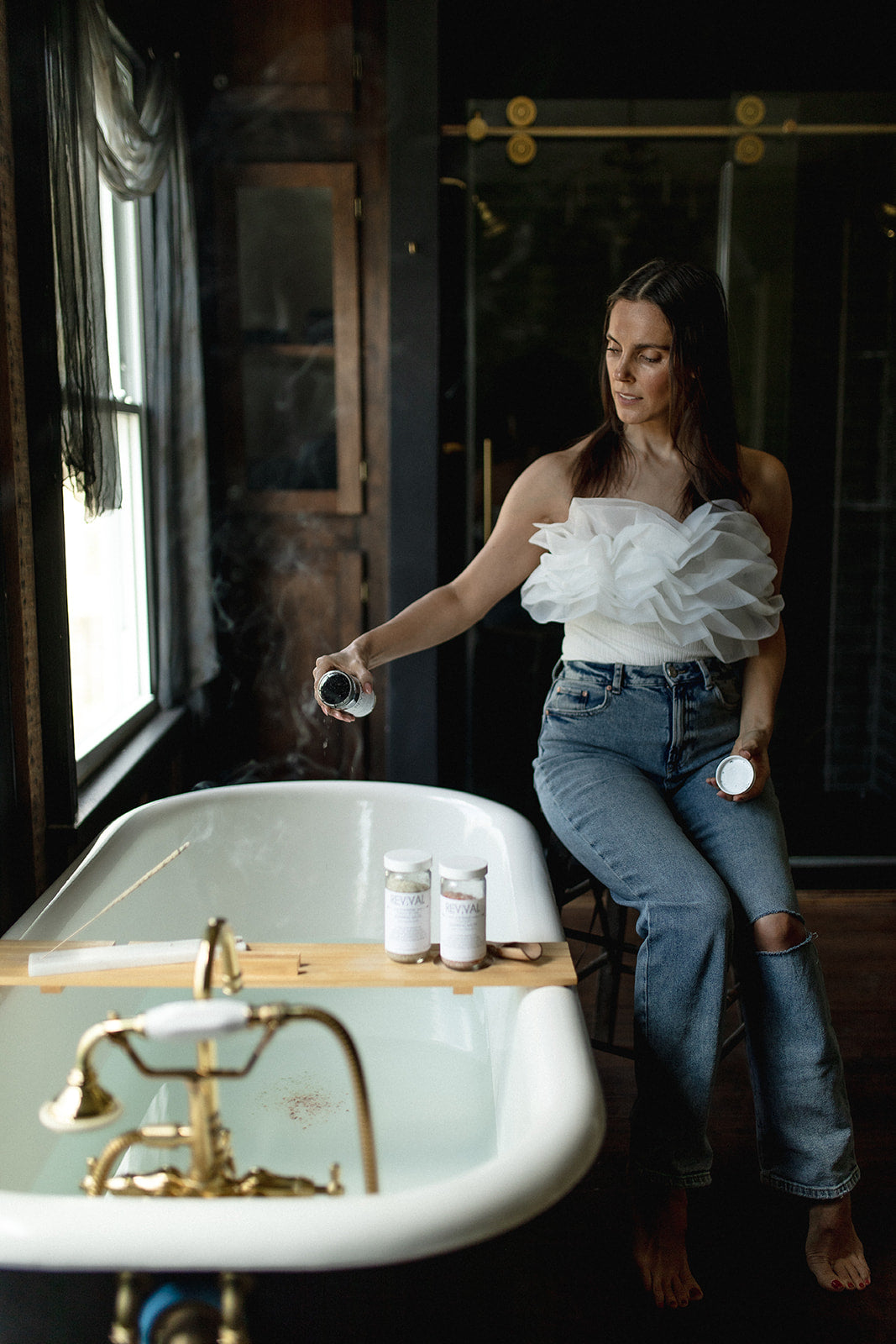 Woman sitting on the edge of a bathtub holding bottles in a dimly lit room.