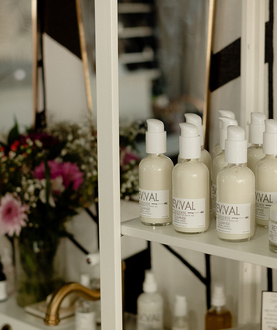 Shelf with skincare products labeled Revival Cleanse Facial Wash in a decorative setting with flowers and a mirror.