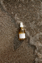 Bottle  of Covet Body Oil labeled 'Revival' on sandy beach with water splash