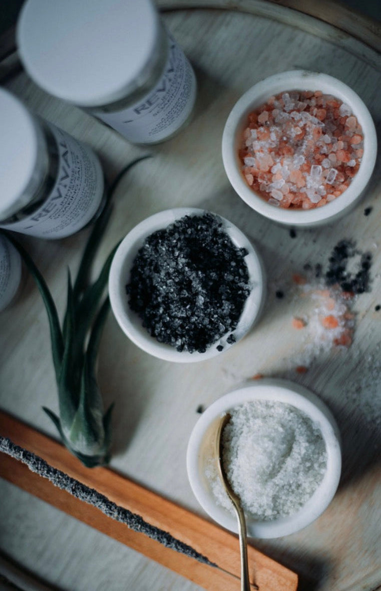 Three small bowls with different types of salt on a wooden surface, with 'Revival' bottles in the background.