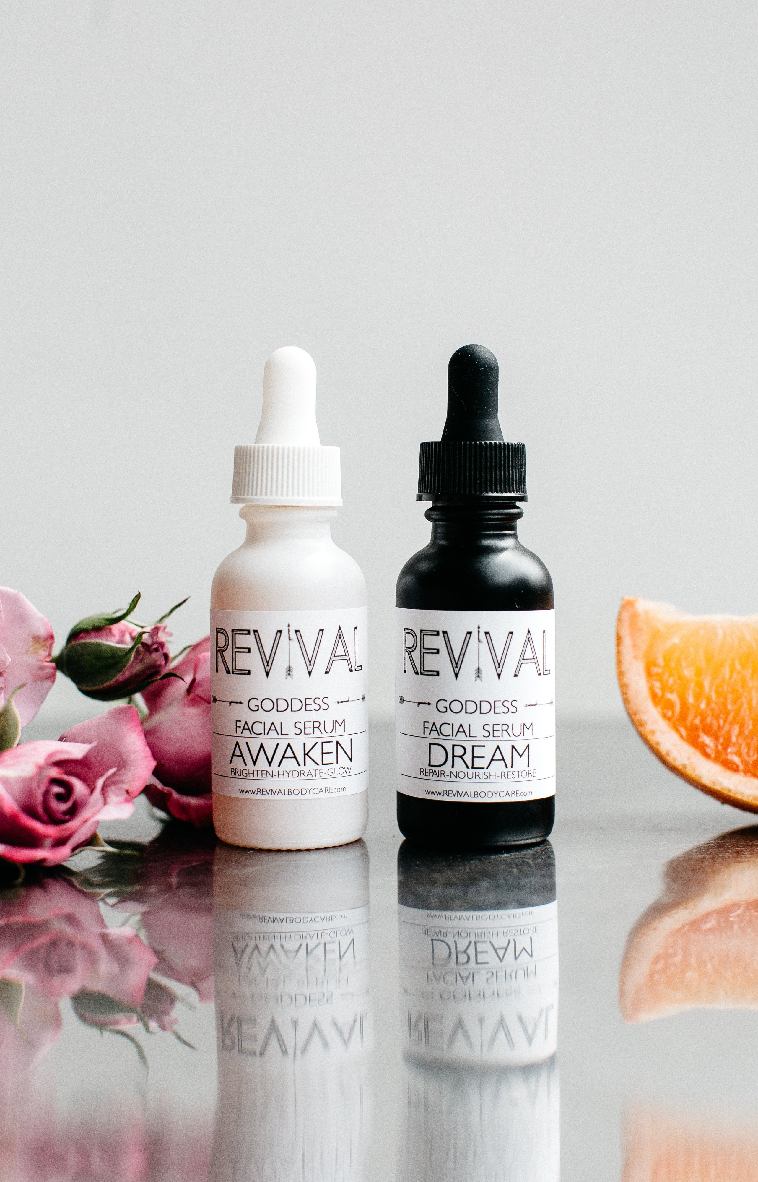 Two bottles of facial serum with dropper caps, labeled 'AWAKEN' and 'DREAM' from the brand REVIVAL. The products are displayed with flowers and a citrus fruit in the background.