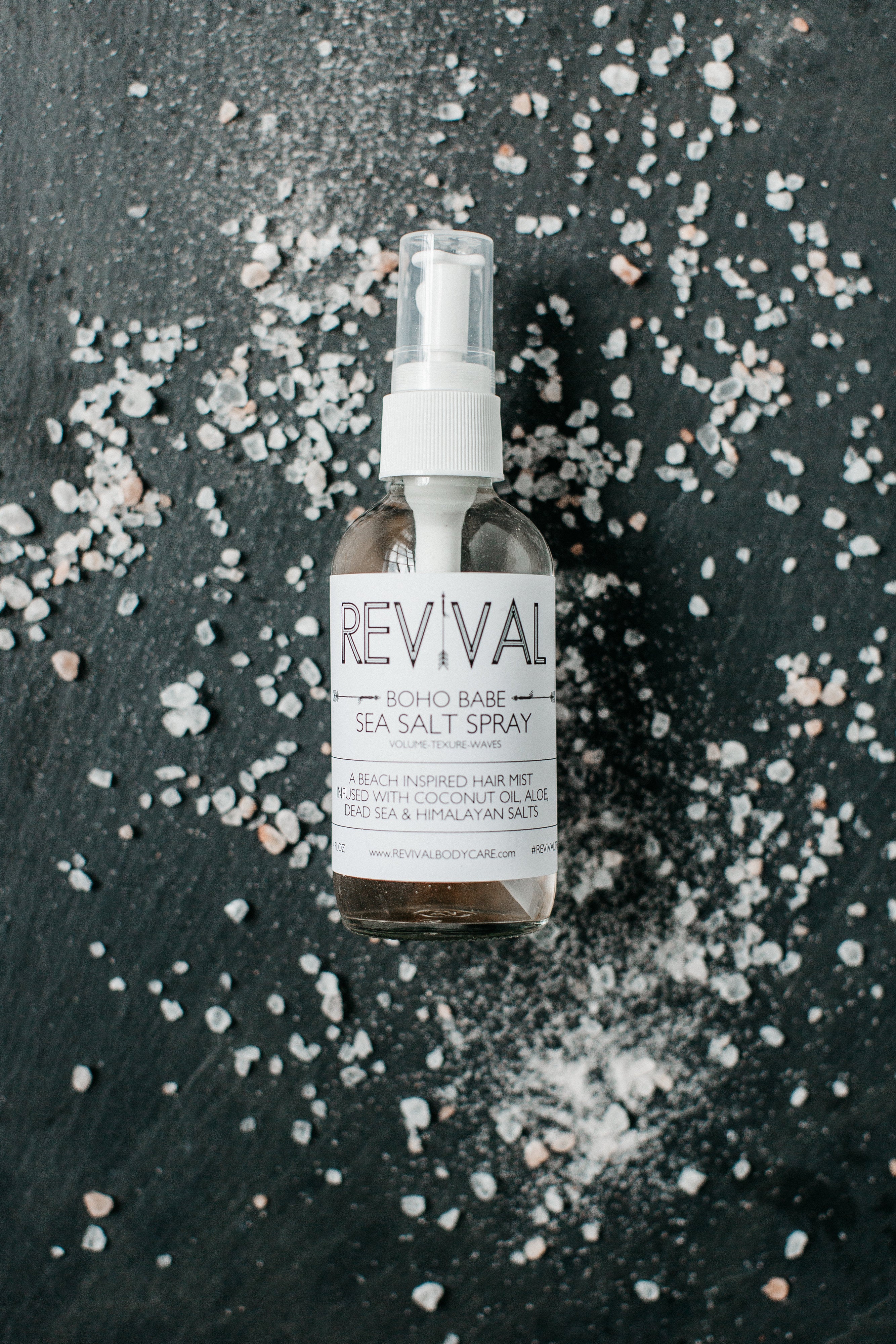 A clear spray bottle labeled 'REVIVAL' with the product name 'BOHO BABE SEA SALT SPRAY' surrounded by scattered sea salt on a dark textured background.
