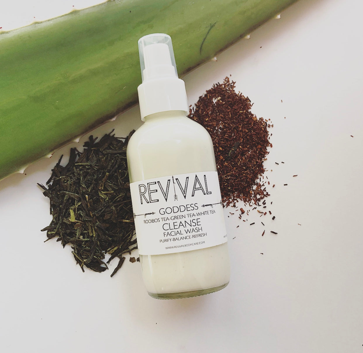A bottle of facial wash labeled 'REVIVAL GODDESS FACIAL WASH - CLEANSE' with a background of aloe vera and loose tea leaves, indicating organic ingredients.