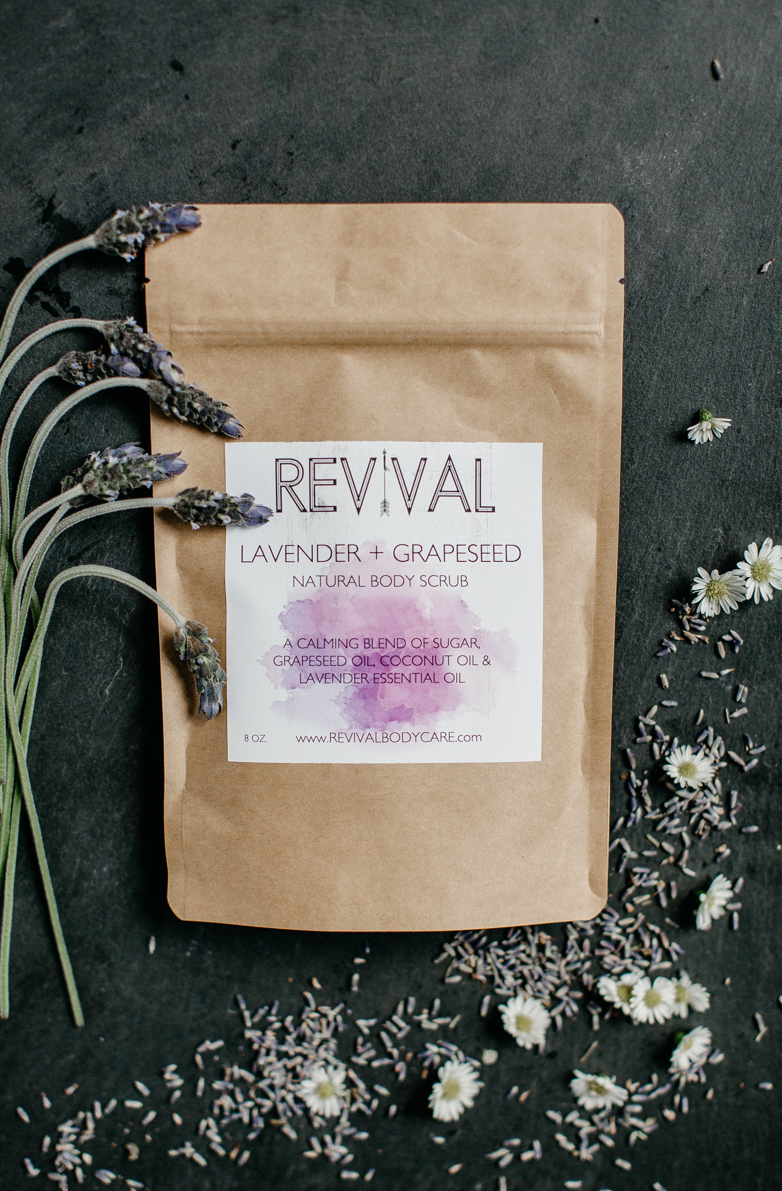 A paper pouch packaging labeled 'REVIVAL' with 'LAVENDER + GRAPESEED' body scrub inside, displayed alongside lavender flowers and scattered grapeseeds on a dark background.
