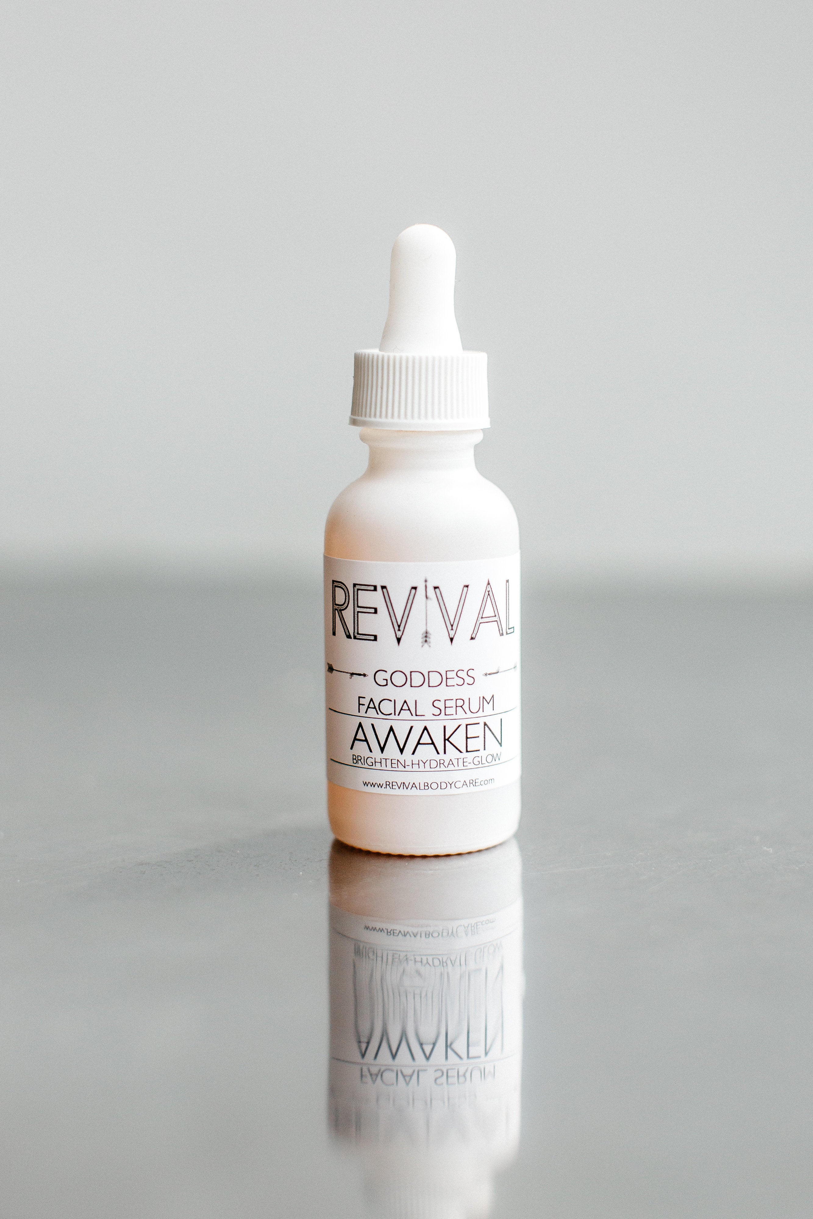 A white bottle with a dropper containing a facial serum, labeled 'REVIVAL GODDESS AWAKEN'. Set against a reflective grey background.
