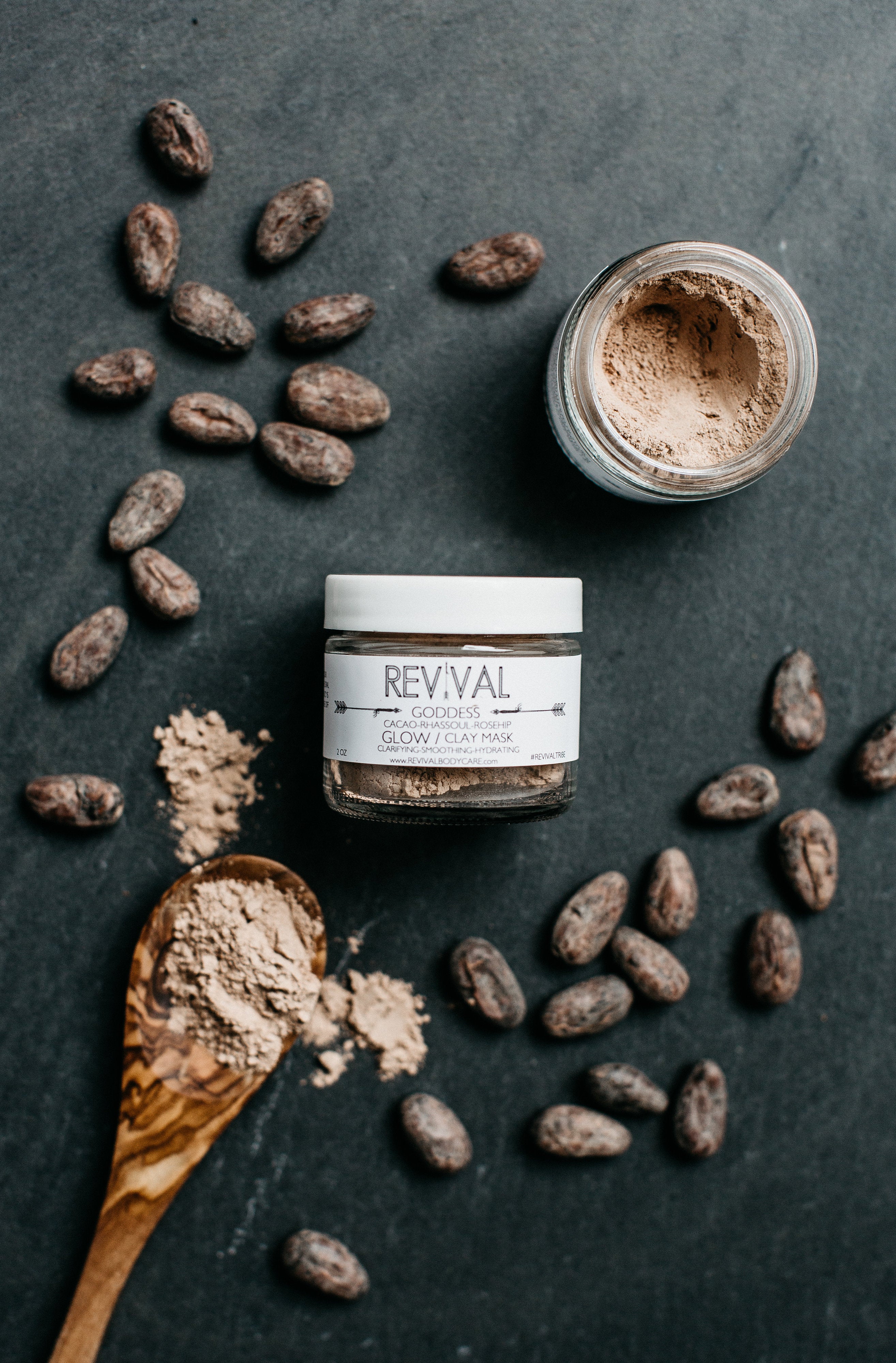A clay mask product by REVIVAL, with natural ingredients like rhassoul clay, cacao, bentonite clay, and rosehips, displayed with a wooden spoon and raw cacao beans on a dark background.