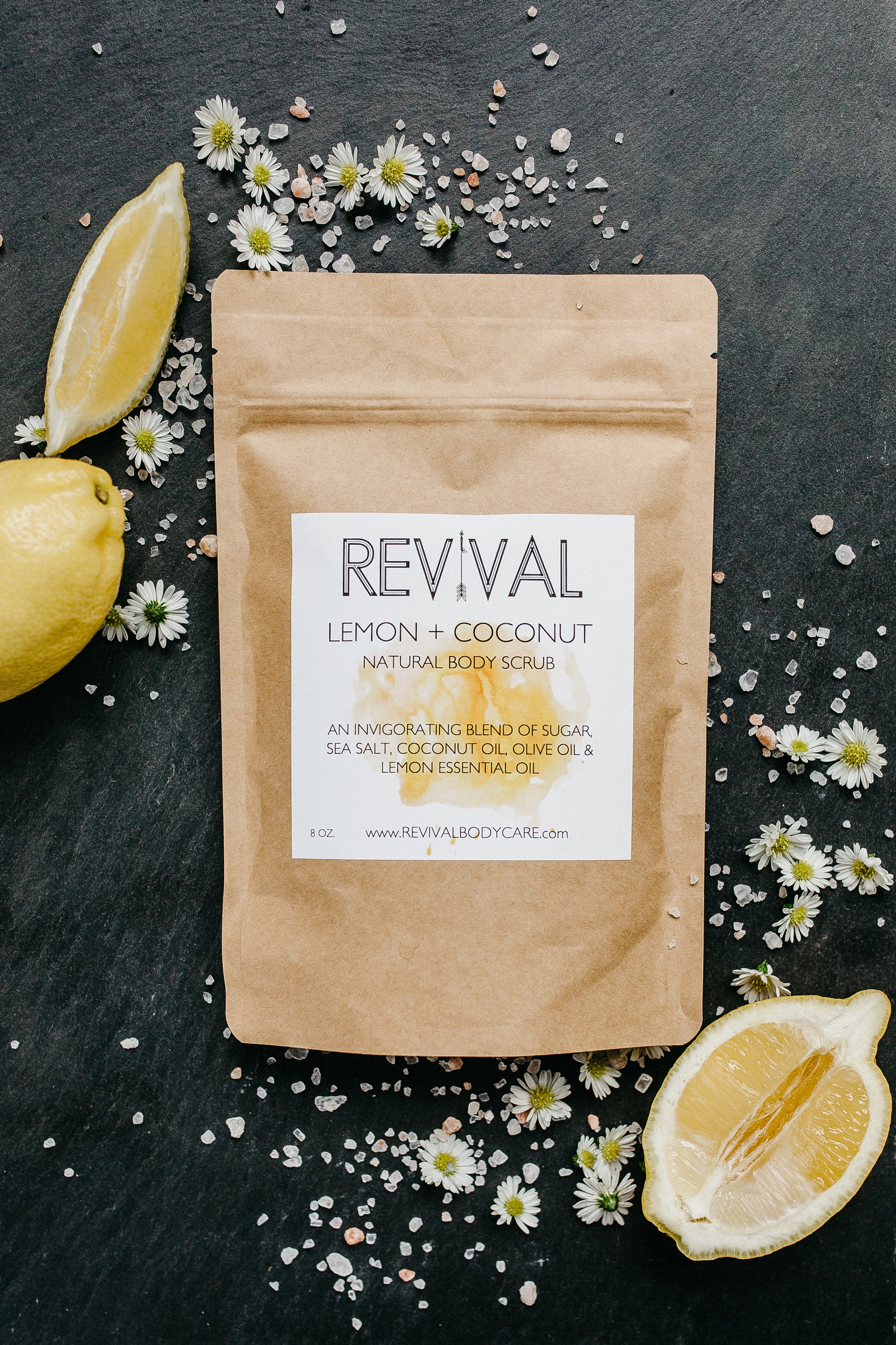 A package of REVIVAL brand Lemon + Coconut body scrub is displayed with lemon slices and scattered flower petals around it on a dark background.