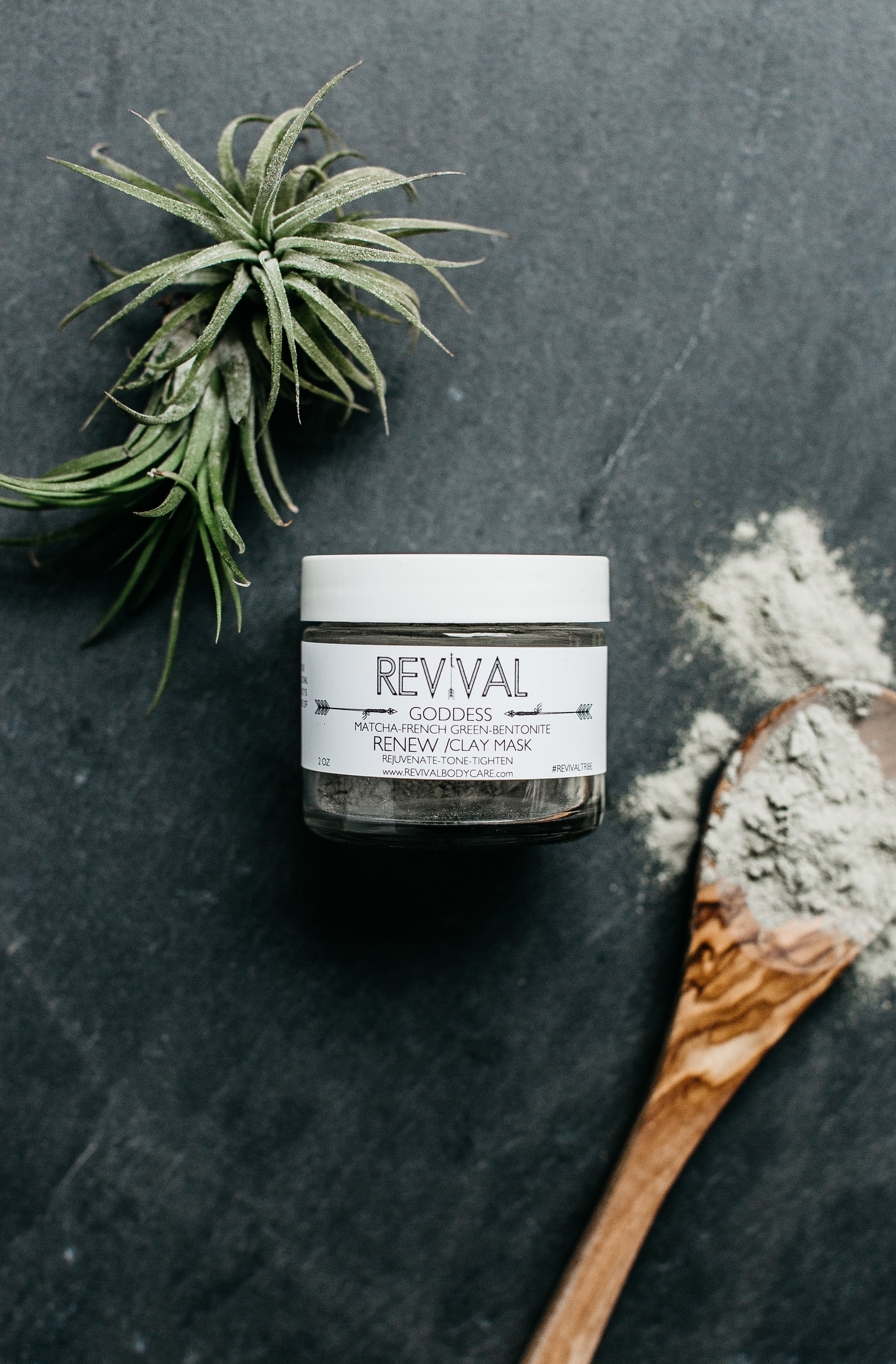 A jar of GODDESS CLAY MASK - RENEW alongside a wooden spoon and a plant, indicating a natural skincare product.