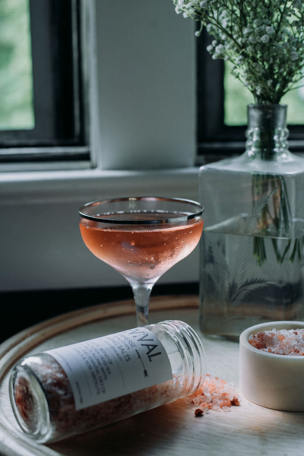 A bottle of IMMERSE soaking salts next to a filled glass on a surface, with a vase of flowers in the background.