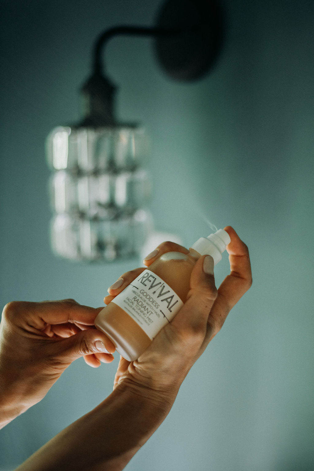 A person's hands holding a bottle of facial toning mist with a soft focus background featuring a hanging light fixture.