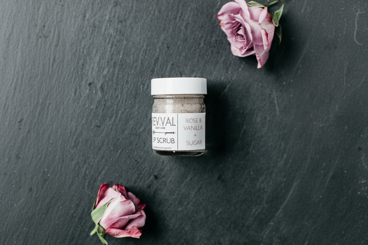 A jar of lip scrub with the label 'ROSE & VANILLA + SUGAR LIP SCRUB' next to two withered pink roses on a dark slate background.