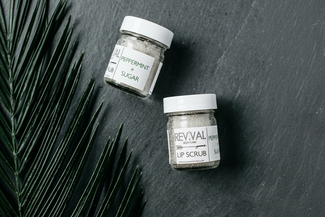 Two small glass jars labeled 'REVIVAL' and 'LIP SCRUB' containing a granular exfoliant, placed on a dark surface with a sprig of mint leaves to the side.